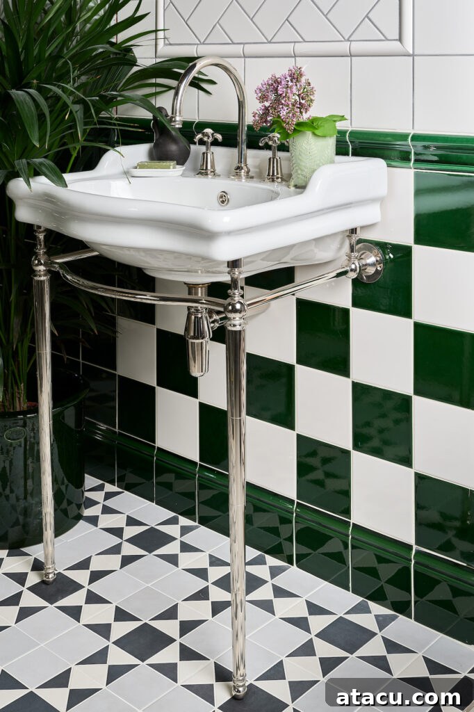 Close-up of a stylish cloakroom featuring a classic green and white chequerboard wall tile pattern, paired with a contrasting geometric black, white, and grey tile floor. A vintage-style white porcelain sink with chrome legs adds elegance, complemented by polished chrome fixtures. Lush greenery and a small floral arrangement bring a touch of nature to the space.