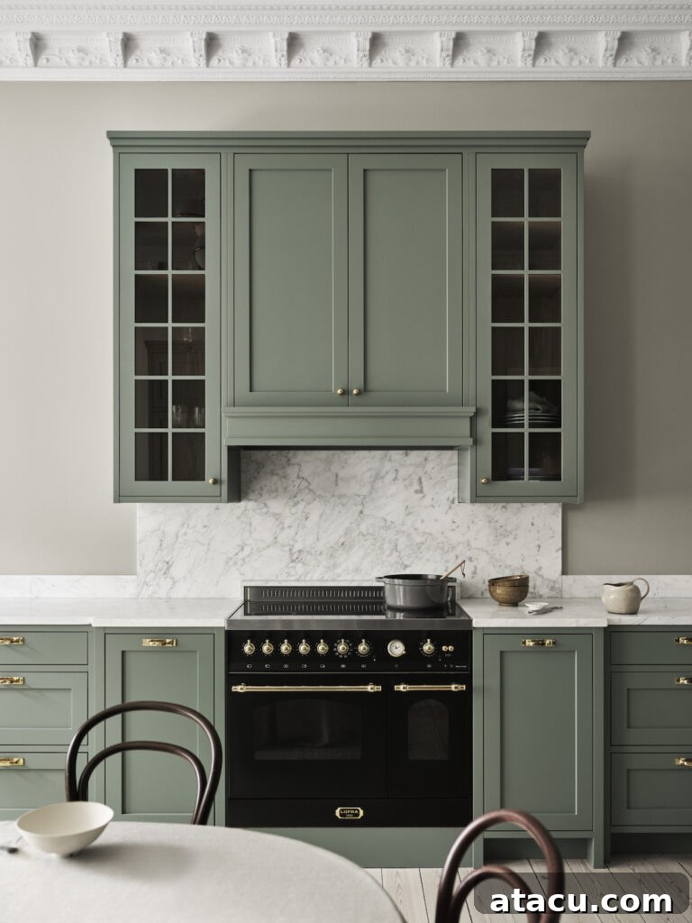 A classic Shaker style kitchen featuring sage green cabinets with brass hardware, a black vintage-style range, marble backsplash and countertop, and a decorative plaster ceiling.