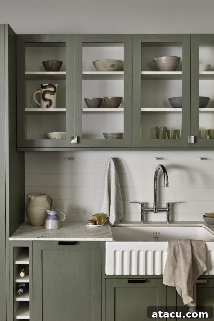 A kitchen with sage green cabinets, glass-paneled upper cupboards displaying ceramic dishes, a farmhouse sink with a chrome faucet, and a marble countertop adorned with neutral-toned accessories.