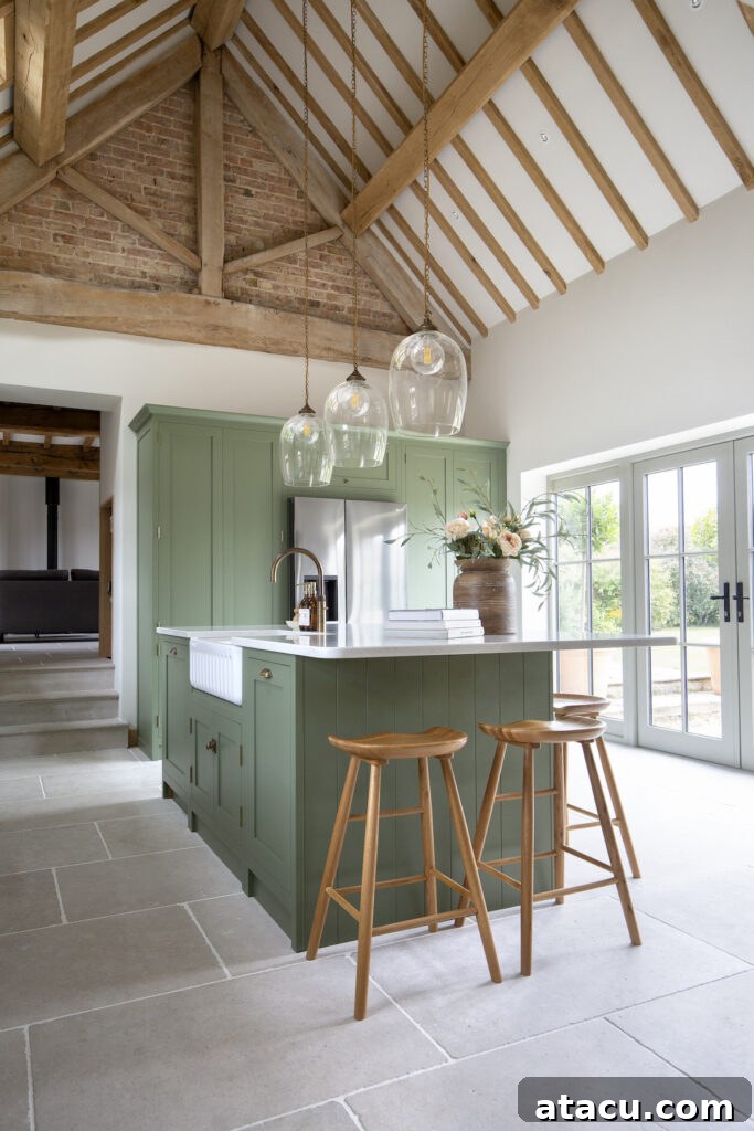 A bright kitchen with sage green cabinetry, a farmhouse sink, wooden bar stools, glass pendant lights, and exposed wooden beams, blending rustic charm with modern elegance.