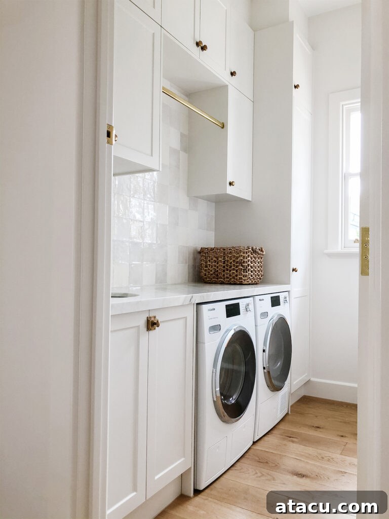 Bright and airy laundry room upstairs featuring white cabinetry with brass accents, a hanging rail, marble countertop, and side-by-side washer and dryer on natural wood flooring.