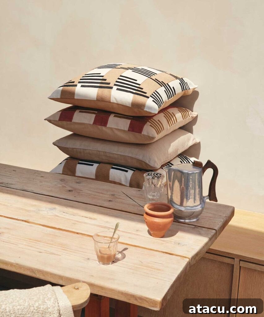 A stack of patterned cushions in earthy tones on a rustic wooden table, surrounded by a teapot, glassware, and clay cup, showcasing luxury home decor accessories.