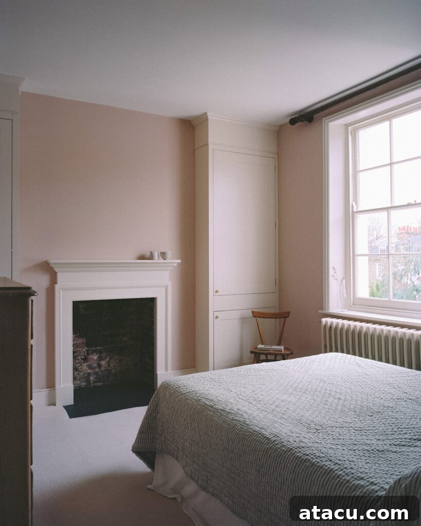A serene bedroom in a Georgian home renovation with soft pink walls, a classic fireplace, and natural light from a large sash window.