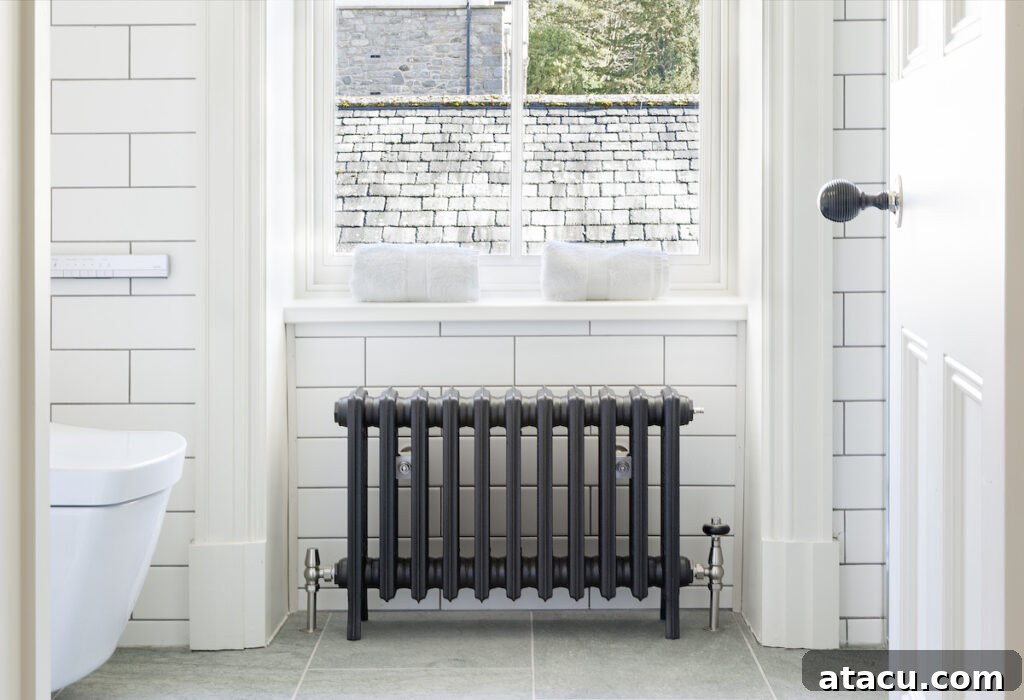 A classic cast iron column radiator positioned beneath a window in a bright, white-tiled bathroom, blending functionality with traditional style.