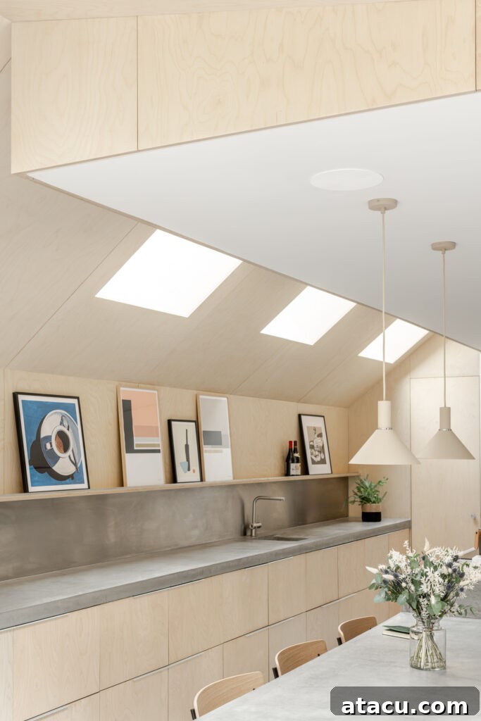 A clean, minimalist kitchen showcasing elegant light wood cabinetry, pristine concrete countertops, and a carefully curated row of framed artwork displayed beneath bright skylights.