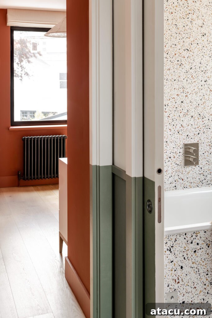 An interior view of a contemporary hallway revealing a warm terracotta-painted room, contrasted by a sleek black radiator, and a glimpse of a stylish terrazzo-tiled bathroom wall.